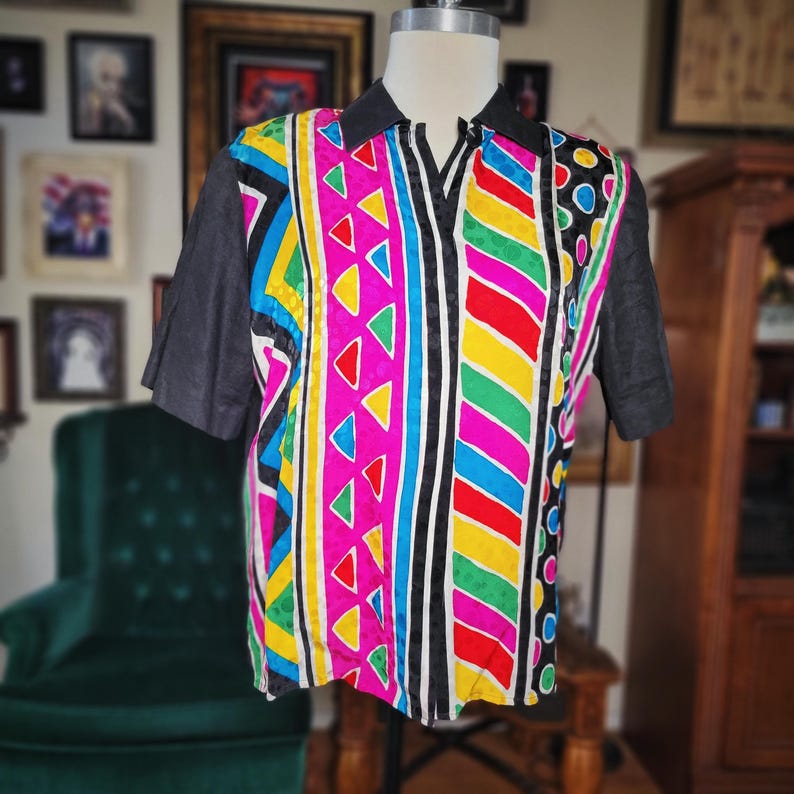 May include: A colorful short-sleeved shirt with a black collar and sleeves. The shirt features a vibrant pattern of vertical stripes in pink, yellow, blue, green, and black, with geometric shapes. The shirt is displayed on a mannequin.