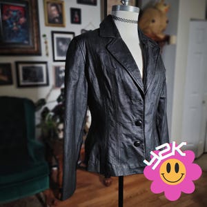 May include: A black leather blazer with a notched collar and two-button closure. The blazer is displayed on a mannequin and has a tailored fit. The image includes a pink flower graphic with the text "Y2K".