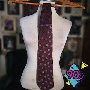 May include: A necktie featuring a deep maroon base color with a repeating pattern of small, light blue squares. The fabric has a subtle, textured appearance, and the tie is displayed hanging straight down from a wooden hanger on a mannequin torso.