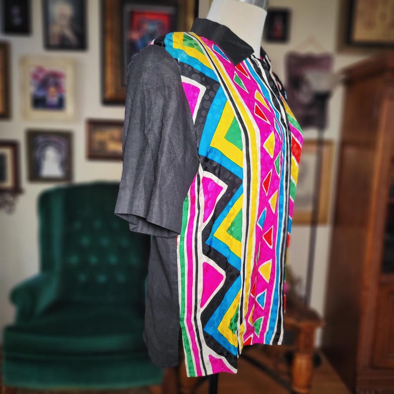 May include: A colorful, patterned shirt with short gray sleeves and a black collar. The shirt's front features a vibrant design with pink, yellow, blue, and green geometric shapes. The shirt is displayed on a mannequin.