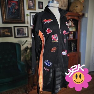 May include: Black zip-up jacket with orange side panels and various embroidered patches. The jacket features the text "meow meow" and other racing-themed patches. The jacket is displayed on a mannequin.
