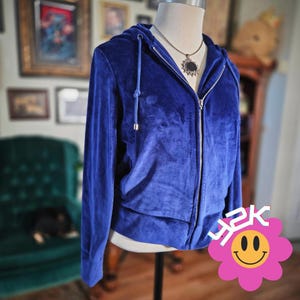 May include: A royal blue velour zip-up hoodie with a drawstring hood and silver zipper. The jacket is displayed on a mannequin and paired with a silver necklace with a black pendant. A pink flower with the text "Y2K" is in the bottom right corner.