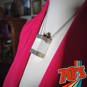 May include: A silver-toned rectangular pendant necklace with a white, rectangular center and ornate silver borders. The necklace hangs on a silver chain against a bright pink jacket. The image has a retro 70's theme.