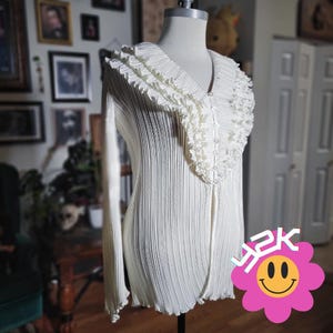 May include: Cream-colored, long-sleeved top with a pleated design and a ruffled collar. The top has a V-neckline and a button-up front. The bottom hem is slightly scalloped. A pink flower with a smiley face and the text "Y2K" is in the bottom right corner.