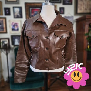 May include: A brown leather jacket with a collared neckline, two front pockets, and button closures. The jacket has long sleeves and lace-up detailing on the sides. The jacket is displayed on a mannequin.
