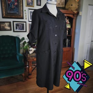 May include: A black button-down dress with short sleeves, two chest pockets, and two hip pockets. The dress is displayed on a mannequin. A choker necklace is visible. The image has a 90s graphic in the bottom right corner.