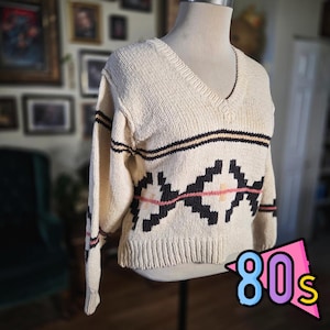 May include: Cream-colored vintage sweater with a V-neck and geometric pattern. The sweater features black, pink, and tan accents. The design includes a diamond-like pattern across the chest and horizontal stripes. The image also includes the text "80s".