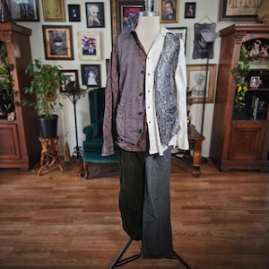 May include: A mannequin displays a split-design outfit. One side features a purple button-up shirt and vest, the other a white shirt with a paisley vest. The pants are split, with one side dark green and the other gray.