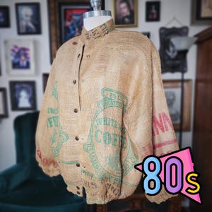 May include: A tan jacket made from a repurposed coffee bean sack. The jacket features a button-up front, a collar, and green and red printed text. The sleeves are gathered at the wrists. The word "80s" is in a pink, blue, and yellow graphic.