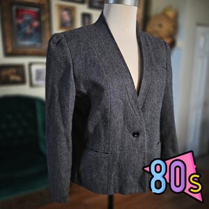 May include: A gray blazer with a notched collar and a single-button closure. The blazer has a tailored fit with shoulder pads and a pocket on the left side. The image includes a colorful "80s" graphic in the bottom right corner.