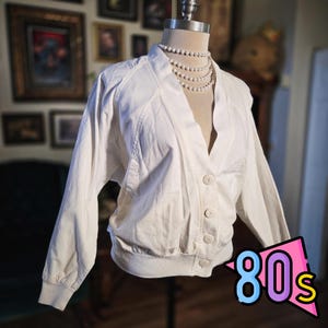 May include: A vintage white jacket with a V-neck, button closure, and ribbed cuffs and hem. The jacket is displayed on a mannequin and paired with a multi-strand pearl necklace. The image includes a colorful "80s" graphic.