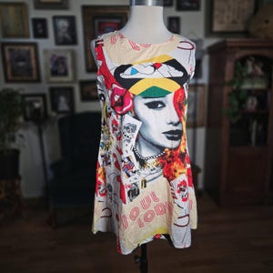 May include: Sleeveless dress with a vibrant graphic print featuring a woman's face, playing cards, and the words "SOUL LODY." The dress has a cream-colored background with red, yellow, and black accents. The dress is displayed on a mannequin.