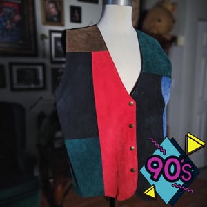 May include: A vintage suede vest with a patchwork design in black, red, green, blue, and brown. The vest has a V-neck and is fastened with gold-colored buttons. The image includes a 90s graphic in the bottom right corner.