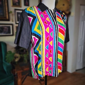 May include: A colorful collared shirt with short sleeves. The shirt features a vibrant geometric pattern in pink, yellow, blue, green, and red. The sleeves and collar are black. The shirt is displayed on a mannequin.