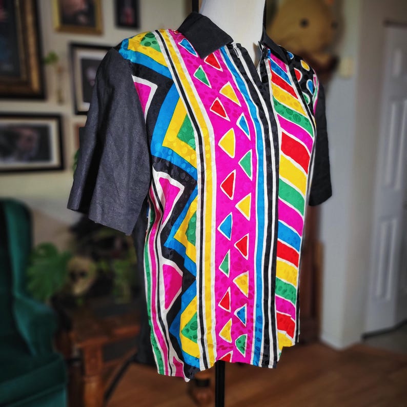 May include: A colorful collared shirt with short sleeves. The shirt features a vibrant geometric pattern in pink, yellow, blue, green, and red. The sleeves and collar are black. The shirt is displayed on a mannequin.