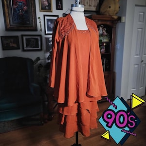 May include: A vintage rust-colored dress set on a mannequin. The set includes a square-neckline dress with a tiered skirt and a matching jacket with intricate lace detailing around the neckline and sleeves. The dress is styled with a black choker.