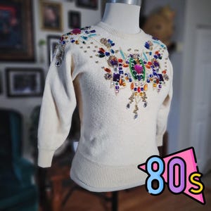 May include: Cream-colored sweater with jewel embellishments. The sweater features a crew neck and long sleeves. The front is adorned with colorful rhinestones and beads in various shapes and sizes, creating a sparkling design. The word "80s" is in a pink and blue graphic.