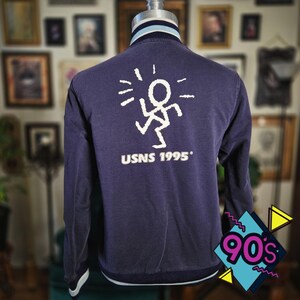 May include: A dark navy blue varsity jacket featuring a white graphic of a stylized dancing stick figure on the back. Below the figure, the text reads USNS 1995. The jacket has a ribbed collar and cuffs with light blue and white striped detailing.