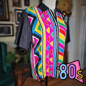 May include: A colorful vintage shirt with a black collar and sleeves. The front features a vibrant geometric pattern in pink, yellow, blue, green, and red. The shirt is labeled with the text "80s" in a retro font.