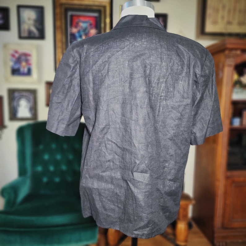 May include: A dark gray short-sleeved button-down shirt. The shirt is made of a textured fabric and has a classic collar. The back of the shirt is visible, showing the cut and fit of the garment. The shirt is displayed on a mannequin.
