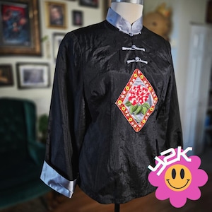 May include: Black long-sleeve shirt with a mandarin collar and frog closures. The shirt features a diamond-shaped embroidered patch with a red flower and green leaves. The cuffs and collar are silver.