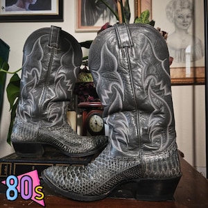 May include: A pair of grey cowboy boots featuring a textured snakeskin-patterned leather foot and a smooth leather shaft with intricate white decorative stitching. The boots have a pointed toe, a stacked heel, and pull tabs at the top of the shaft.