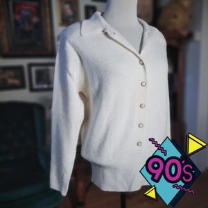 May include: Cream-colored cardigan sweater with a collared neckline and button-down front. The long-sleeved sweater has a ribbed hem and cuffs. The image includes a 90s graphic in the bottom right corner.