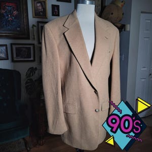 May include: A tan blazer displayed on a mannequin. The blazer has a notched lapel, two front pockets, and a single-button closure. The blazer is made of a textured fabric and has a classic, tailored fit. The image has a 90s graphic in the bottom right corner.