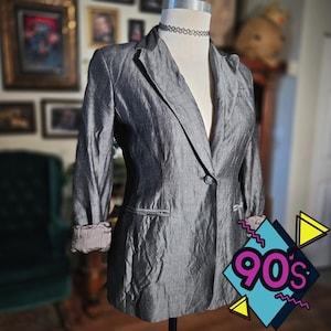 May include: A gray blazer with a single button closure and a notched lapel. The sleeves are rolled up, revealing a contrasting lining. A black choker necklace is around the mannequin's neck. The image has a 90's graphic in the bottom right corner.