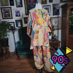 May include: A vintage two-piece outfit with a cream background and a colorful paisley pattern. The top has a mandarin collar and long sleeves. The pants are loose-fitting. The outfit is displayed on a mannequin.