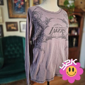 May include: A mauve long-sleeve thermal shirt featuring a black graphic print of the Los Angeles Lakers logo and basketball, surrounded by intricate swirling scrollwork designs. The shirt has a crew neckline and visible black stitching details.