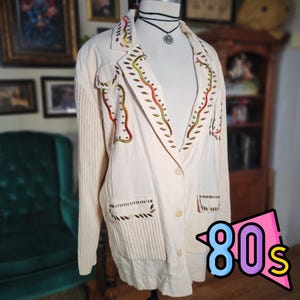 May include: Cream-colored blazer with colorful embroidered details. The blazer has a ribbed knit sleeve and pocket design. The text "80s" is in a pink, blue, and yellow graphic. The blazer is buttoned up.
