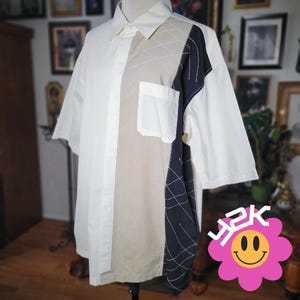 May include: A short-sleeved button-down shirt with a unique color-block design. The shirt features a white, beige, and navy blue color scheme. The beige section has a grid pattern, and the navy blue section has a white grid pattern. A white pocket is on the chest.