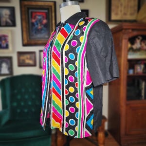May include: A colorful, patterned blouse with short sleeves. The front features vertical stripes of red, yellow, blue, and green, with a black polka dot design. The sleeves and collar are solid black.