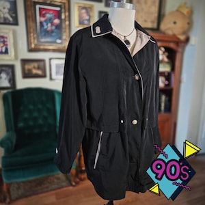 May include: A black jacket with a beige lining and white trim. The jacket has a collared neckline, gold buttons, and a drawstring waist. The jacket is displayed on a mannequin. The background includes a green chair and framed pictures.