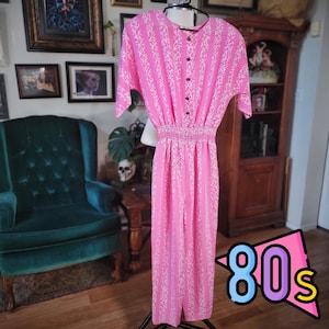 May include: A pink jumpsuit with a white patterned design, short sleeves, and a button-up front. The jumpsuit has a cinched waist and wide legs. The image has a retro 80s theme.