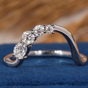 May include: A curved, silver-toned ring with five clear, round-cut gemstones. The ring is set against a blue velvet surface, with a blurred brown background.