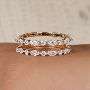 Könnte beinhalten: Ein doppelreihiger Goldring mit klaren, marquiseförmigen und runden Edelsteinen. Der Ring wird auf einer Hand gezeigt, wobei das detaillierte Design und das Funkeln der Steine hervorgehoben werden. Die Ringbänder verlaufen parallel.