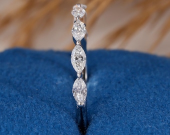 May include: A delicate silver ring featuring four marquise-cut diamonds and small round diamonds. The ring is set on a blue surface, with a blurred background of dried plants.