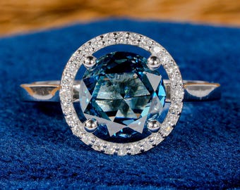 May include: A silver ring with a large, teal-coloured, faceted gemstone encircled by a halo of smaller, clear stones. The ring is set on a deep blue fabric, providing a striking contrast.
