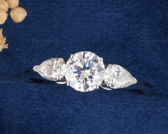 May include: A three-stone engagement ring with a large round center diamond and two pear-shaped side diamonds set in a white gold band. The ring is displayed in a blue velvet ring box.