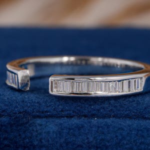 Op de afbeelding: Een zilveren ring met een rij rechthoekig geslepen diamanten. De ring heeft een open ontwerp met een opening in de band. De ring wordt gepresenteerd op een blauw fluwelen oppervlak.