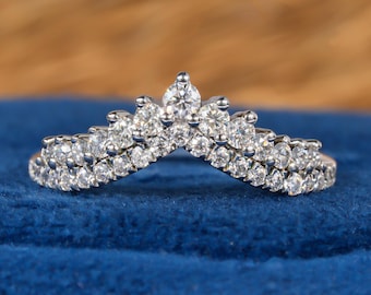 May include: A silver-toned, V-shaped ring with numerous small, clear gemstones. The ring's design features a central, pear-shaped stone, with smaller stones arranged in rows along the band. The ring is set against a blue velvet surface.