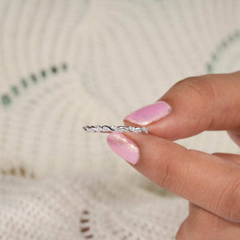 Pu&ograve; includere: Un delicato anello d'argento con un design ondulato, adornato con piccole pietre preziose scintillanti. L'anello &egrave; tenuto tra due dita, mostrando i suoi dettagli intricati e il suo stile elegante. L'anello sembra essere una fede nuziale o un anello di anniversario.