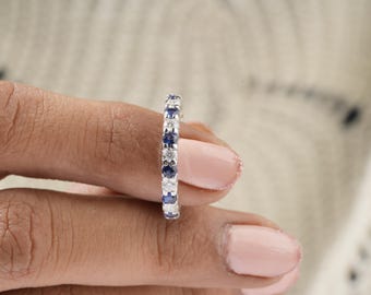 May include: A close-up of a ring featuring alternating blue and clear gemstones set in a silver band. The ring is held between fingers with pink nail polish, against a blurred background.