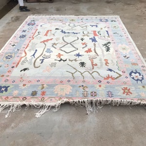 May include: A large, rectangular rug with a light blue field and a border in pink and blue. The rug features a variety of colorful, abstract shapes and designs, including floral and geometric patterns. The edges have a fringe.