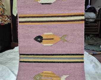 May include: A rectangular, hand-woven rug with a pink background. The rug features three horizontal stripes with alternating colors of black, yellow, and brown. It has four fish motifs in shades of yellow, white, and black.