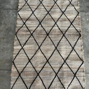 May include: A beige and black diamond patterned rug with a white fringe border. The rug is made of a woven material and has a simple, modern design.