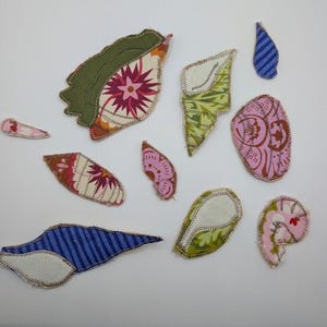 May include: A collection of fabric leaf shapes in various sizes and patterns. The leaves feature a variety of colors, including green, pink, blue, and white, with floral and striped designs. Each leaf is outlined with a contrasting stitch.