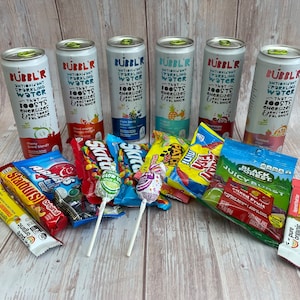 May include: A variety of colorful candies and lollipops, including Skittles, Starburst, and Juicy Burst, are arranged on a wooden surface. Six cans of Bubbl'R Antioxidant Sparkling Water are also visible, each with a different flavor: Cherry Citrus Blend, Blood Orange Mango, Triple Berry Sweet, Grape, Organic, and Twisted.